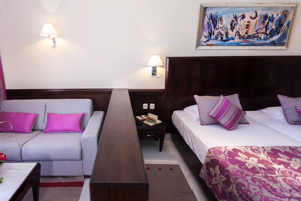 A hotel room with a grey sofa, two beds with purple and white bedding, a wooden headboard, a painting on the wall, and a nightstand with a lamp.
