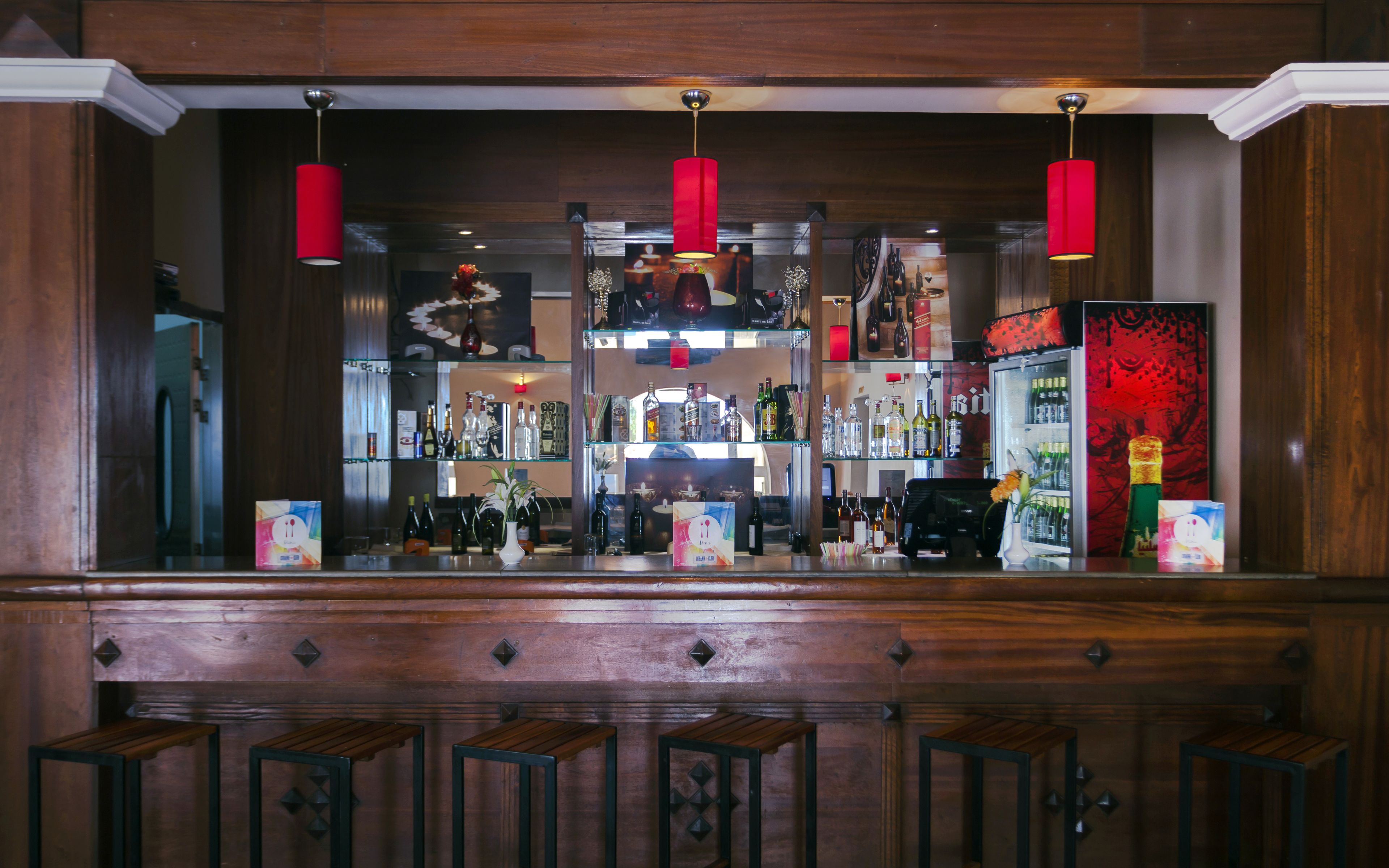 A bar area with wooden paneling, red pendant lights, and shelves stocked with bottles.