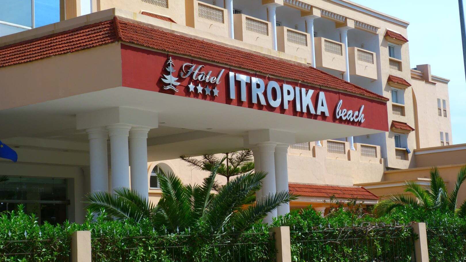 Un hotel llamado "Hotel Tropika Beach" con fachada roja y blanca, columnas blancas y un área de balcón.