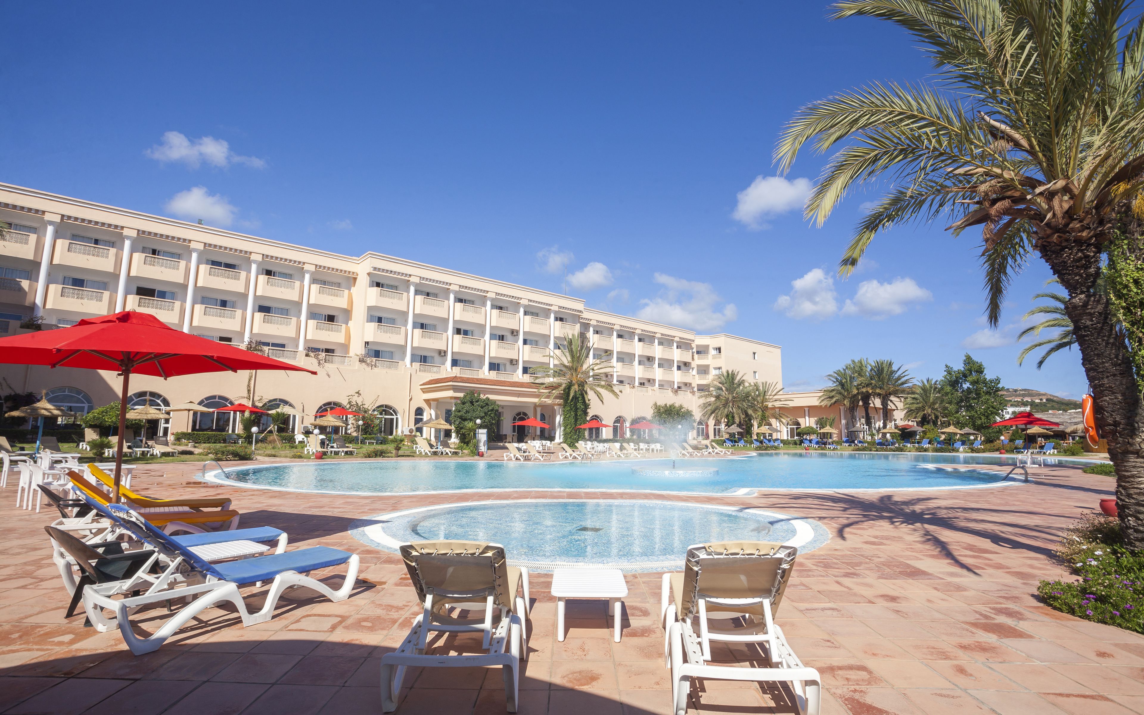 A hotel pool area with sun loungers, umbrellas, and a large building in the background.