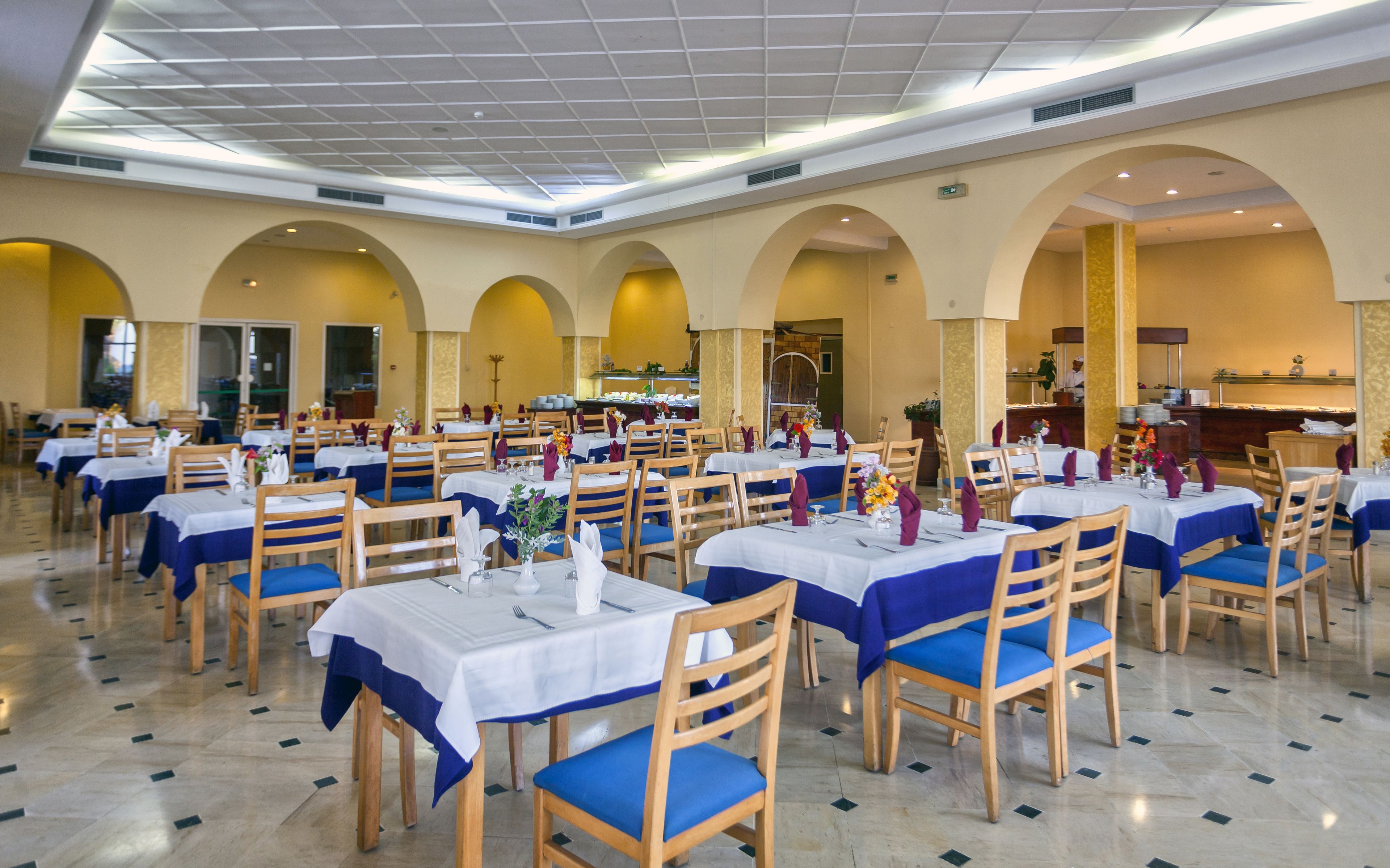 A spacious dining area with multiple tables and chairs, featuring a high ceiling with skylights and a central kitchen area.