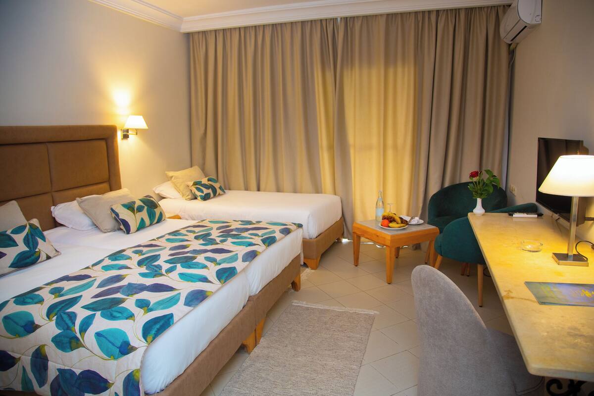 A hotel room with two beds, a desk, a chair, a lamp, and a TV.