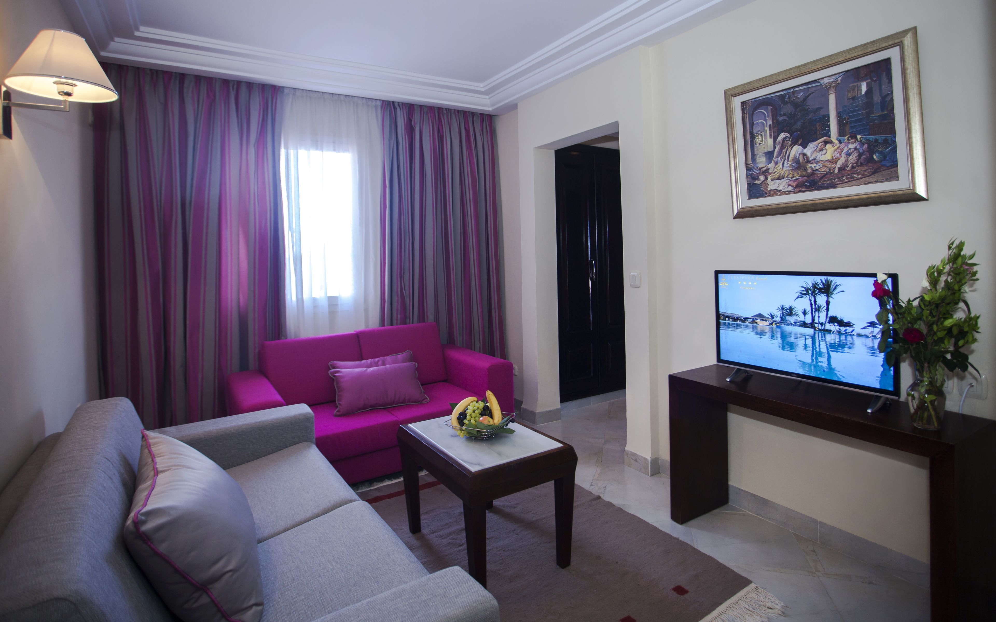 A living room with a pink sofa, a grey sofa, a coffee table with fruit, a TV on a stand, and a painting on the wall.