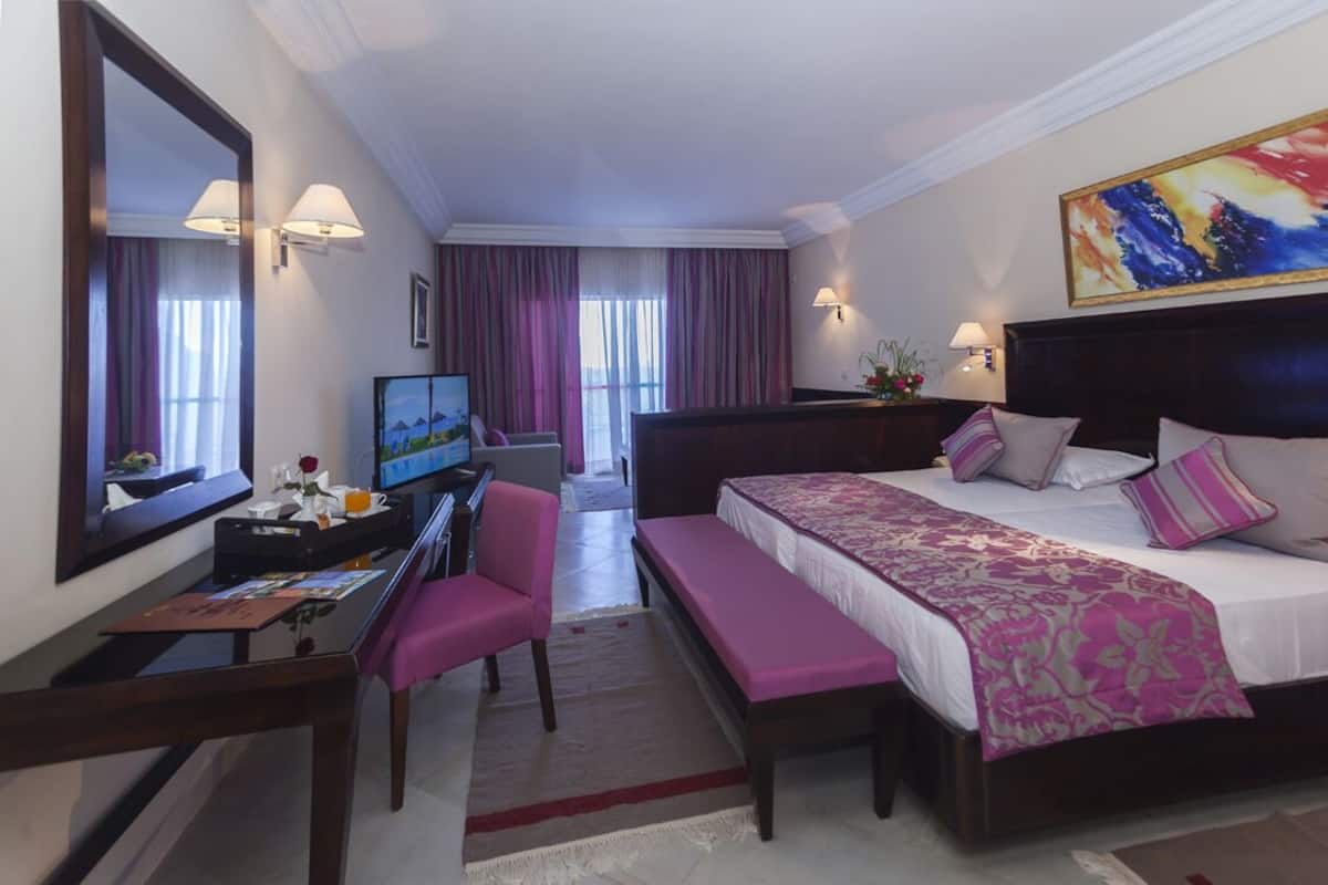 A hotel room with a large bed, a desk with a computer, a chair, a bench, and a television.