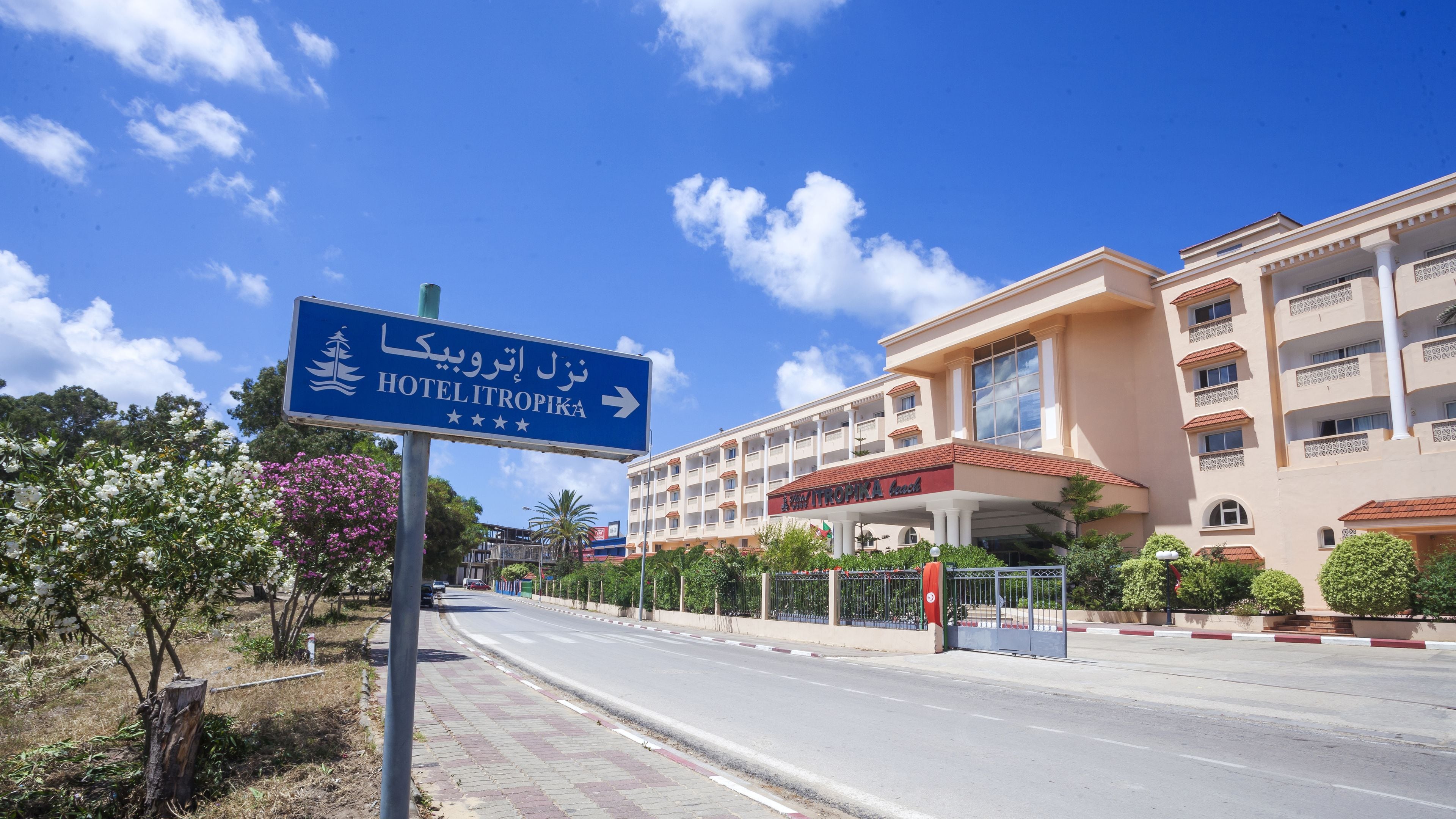 Entrada de hotel com uma placa para Hotelitropika, um edifício com vários andares e telhado vermelho, sob um céu azul límpido.