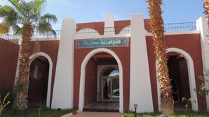 A building entrance with a sign in Arabic, palm trees, and a clear sky. - Hôtel Gourara (Timimoun)