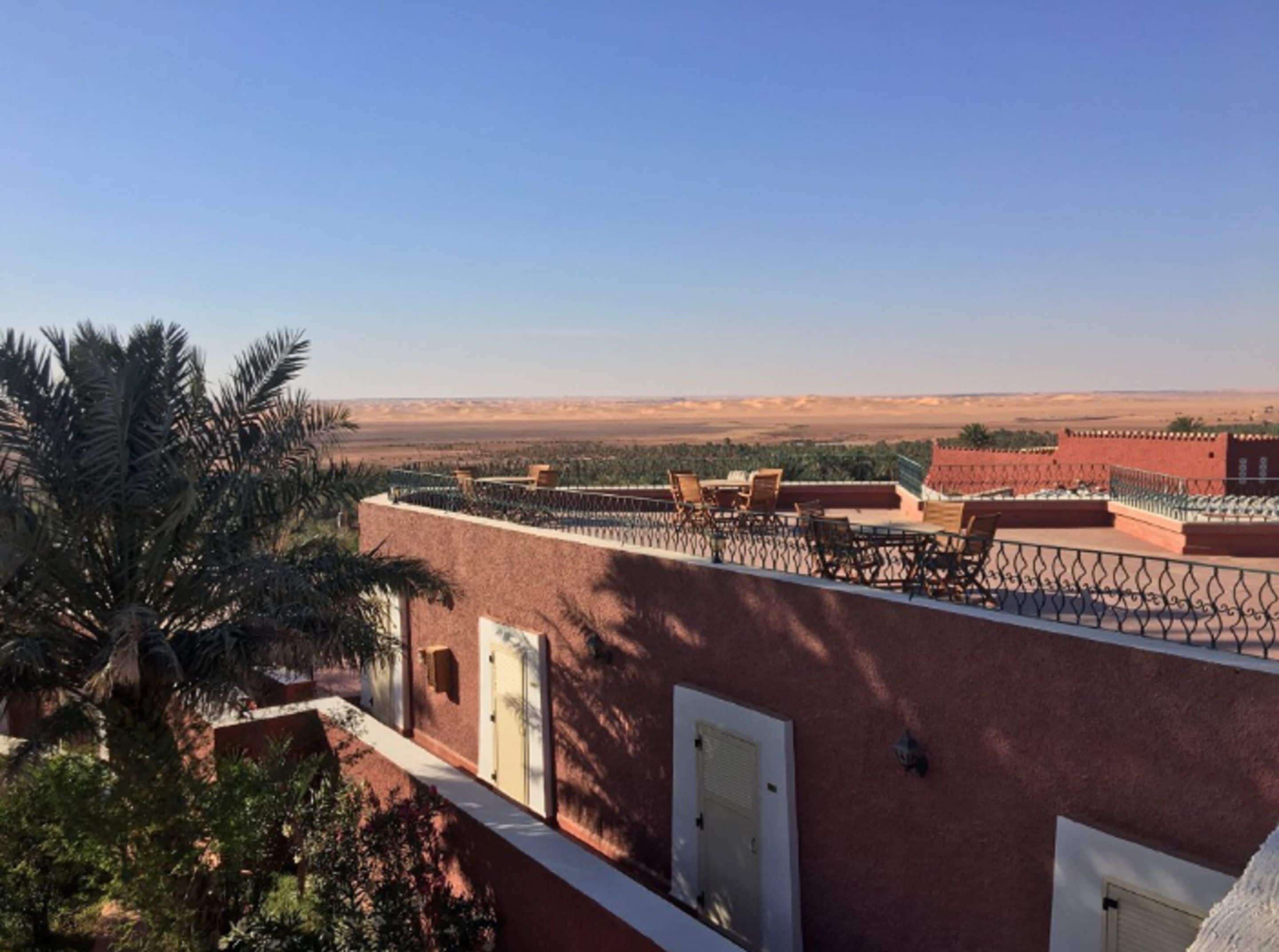 Une terrasse sur le toit offrant une vue sur un paysage désertique, des palmiers et un bâtiment en briques rouges.