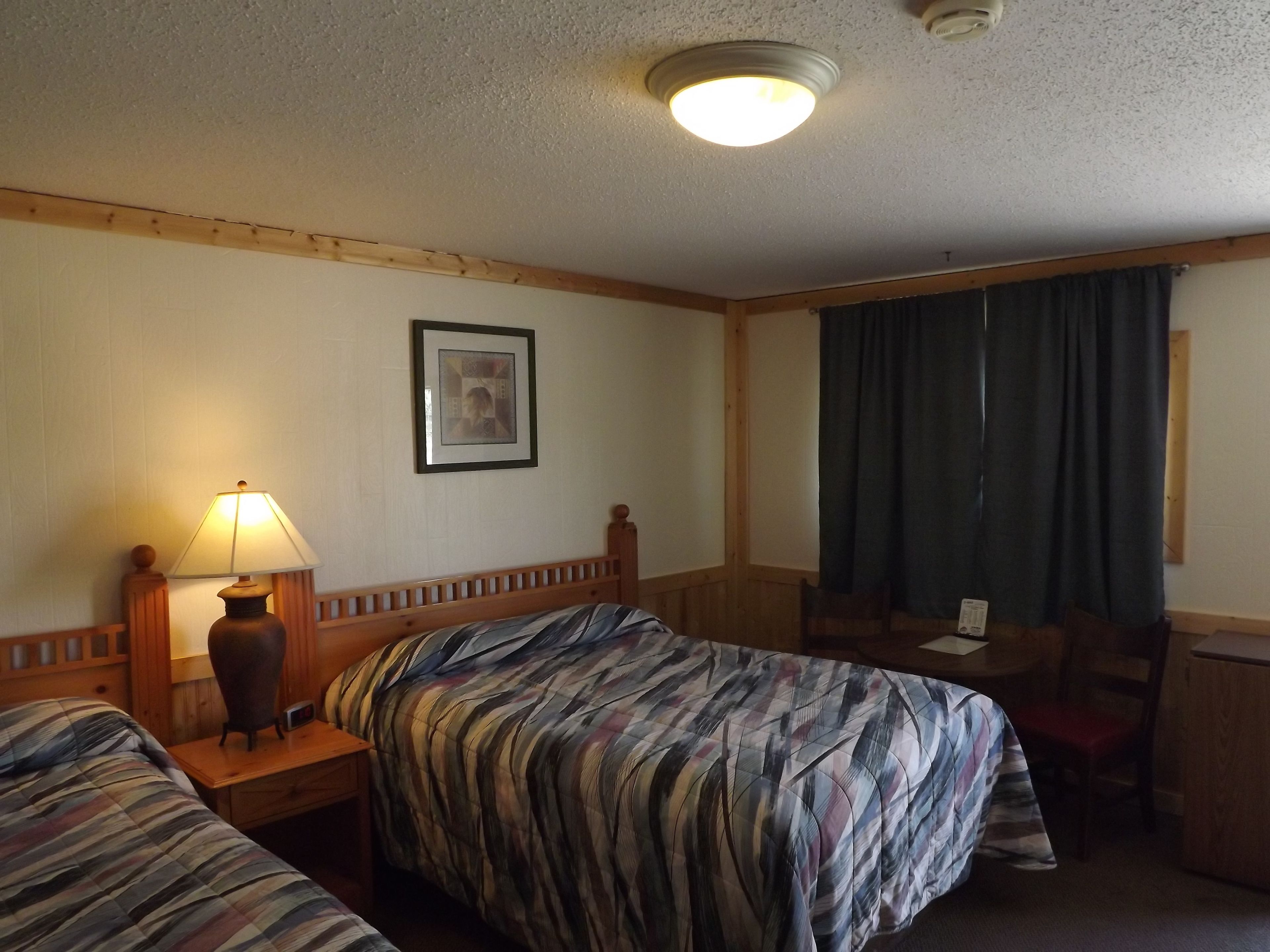 A hotel room with two beds, a desk, a chair, a lamp, a framed picture on the wall, and a window with curtains.
