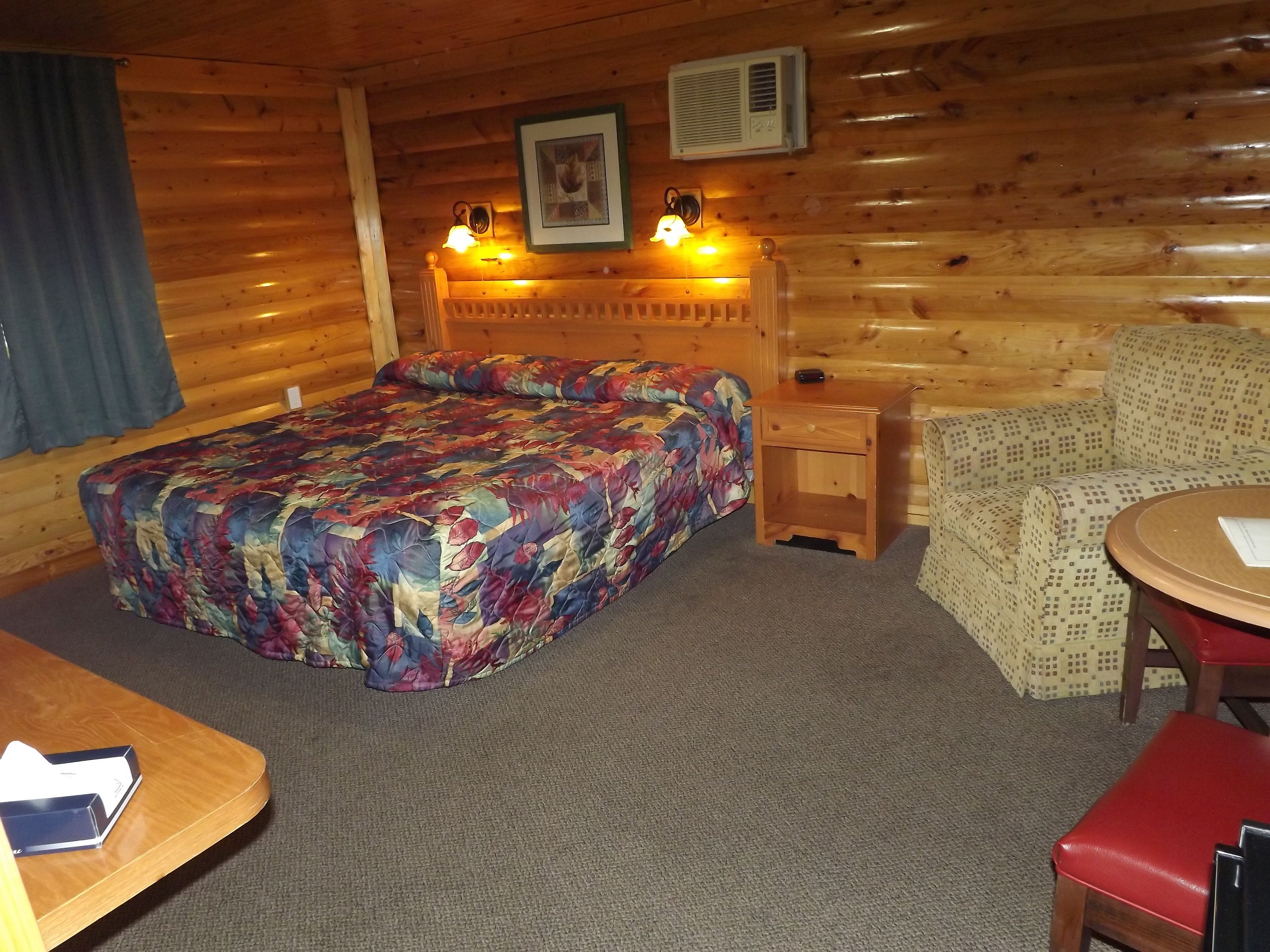 A cozy wooden cabin room with a bed, a small table, a chair, and a lamp.