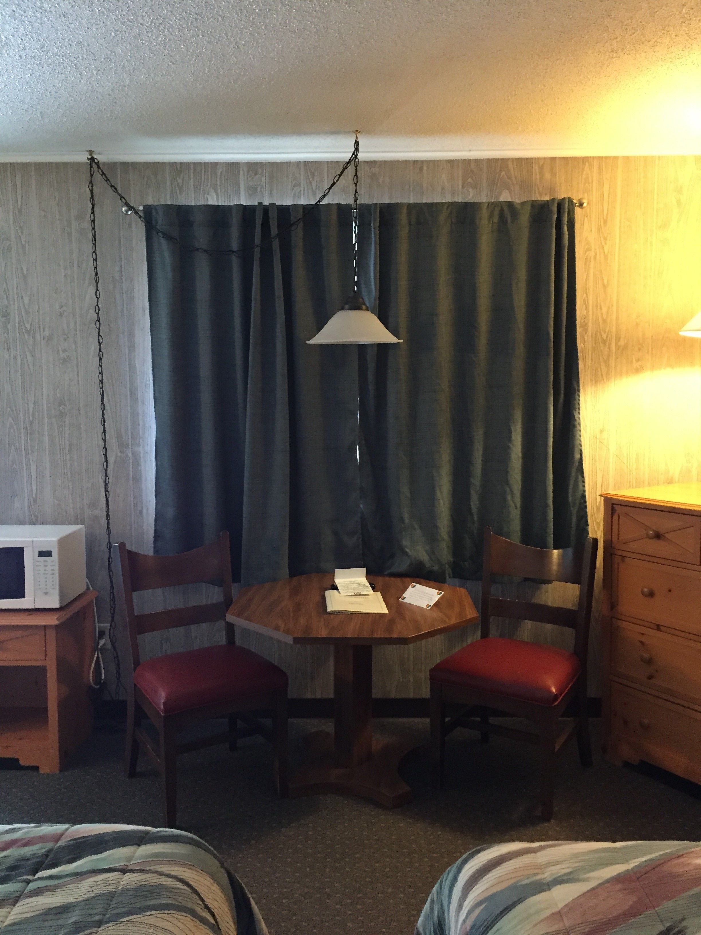 Habitación de hotel con una mesa de madera, dos sillas, microondas, cómoda y lámpara colgante.