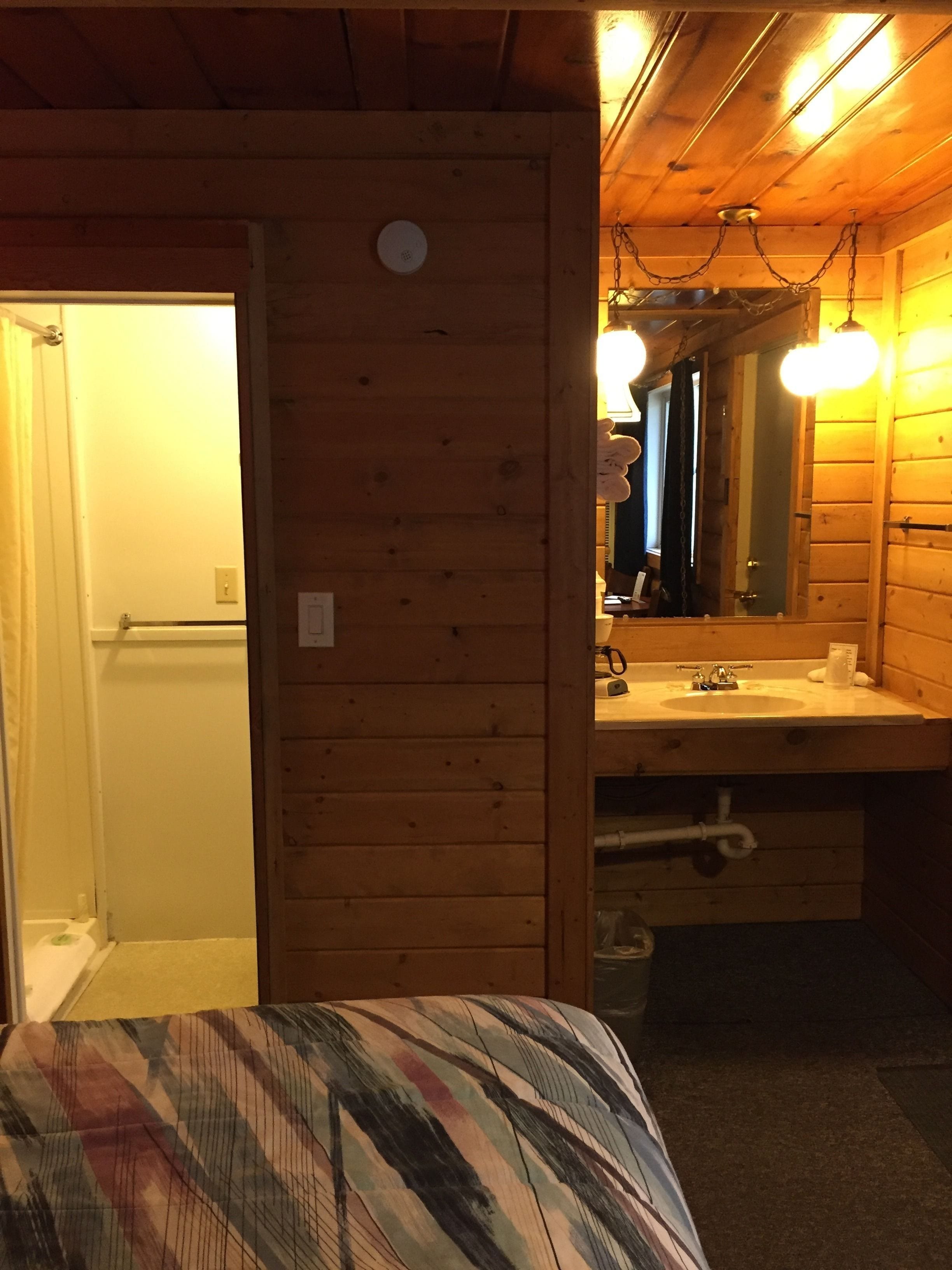 A wooden cabin interior with a bed, a bathroom with a sink and mirror, and a door leading to another room.