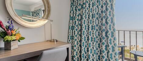 A hotel room with a wooden desk, a chair, a round mirror, and a view of the sea through patterned curtains.