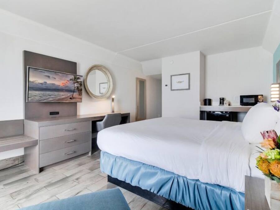 Habitació d'hotel amb un llit, una taula de treball, una cadira, un televisor, un mirall i un arranjament floral.