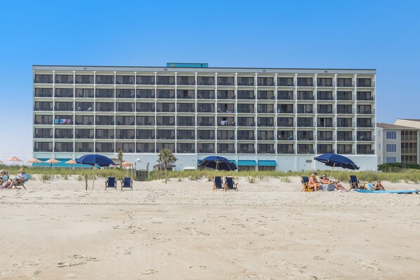 Un hôtel à plusieurs étages, à l’architecture extérieure uniforme, avec une plage au premier plan, des chaises de plage et des parasols.