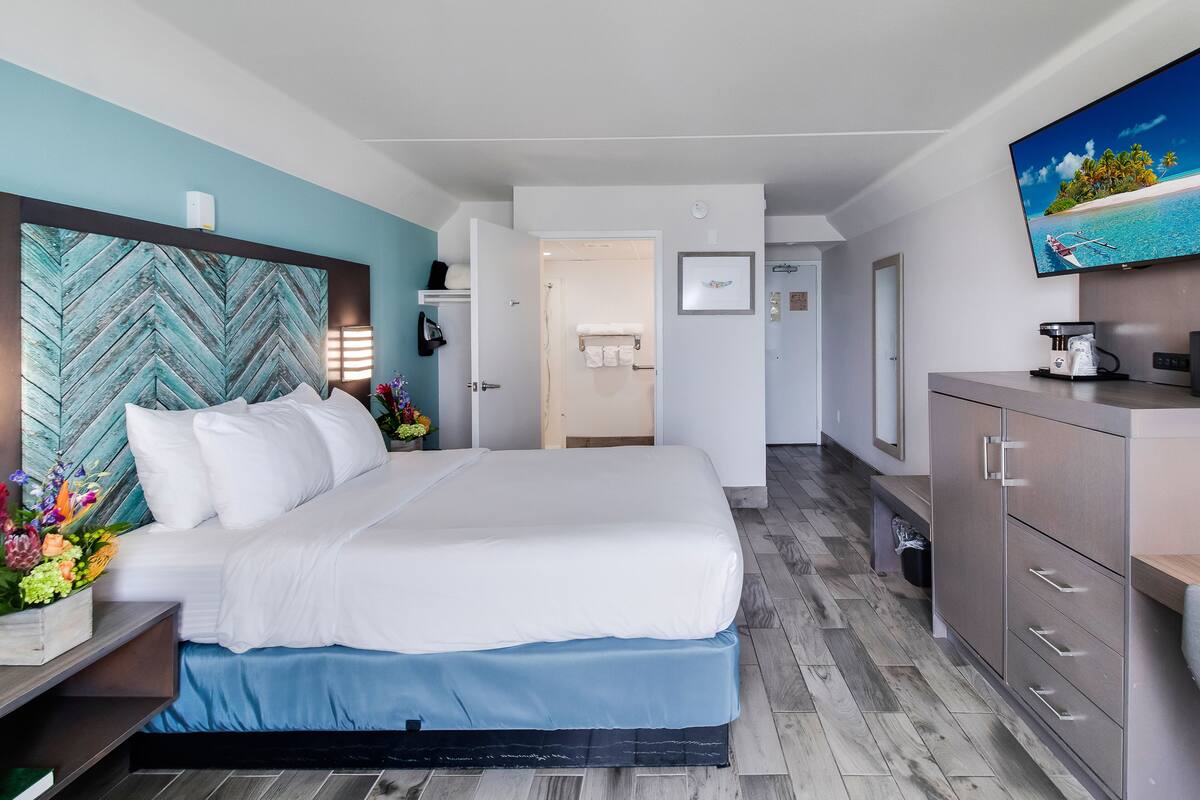 Ein Hotelzimmer mit Bett, Schreibtisch, Kommode und Fernseher, der eine tropische Szene zeigt.