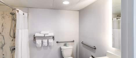A modern bathroom with a white toilet, shower, and sink. There are towels on a rack and a handrail for accessibility.