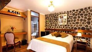 A hotel room with a large bed, a wooden headboard, a stone wall, a small desk with a chair, and a bathroom visible through an open door. - El Portal de San Lázaro (Arequipa)