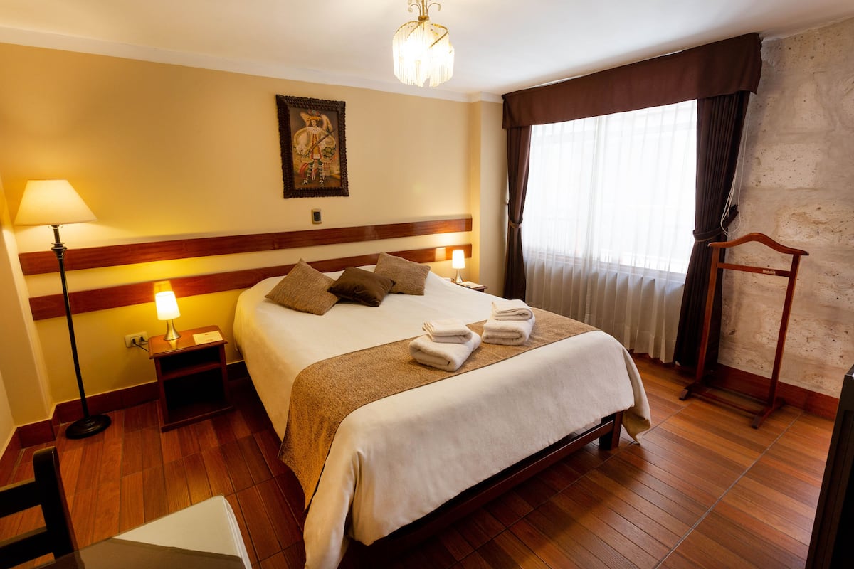 Een hotelkamer met een groot bed, houten vloer, een nachtkastje met lamp, een schilderij aan de muur en een raam met gordijnen.
