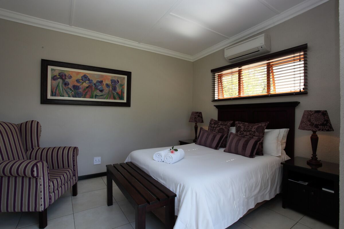 Habitación de hotel con cama, una silla, un banco, una mesita de noche, una lámpara, un cuadro y una ventana con persianas.