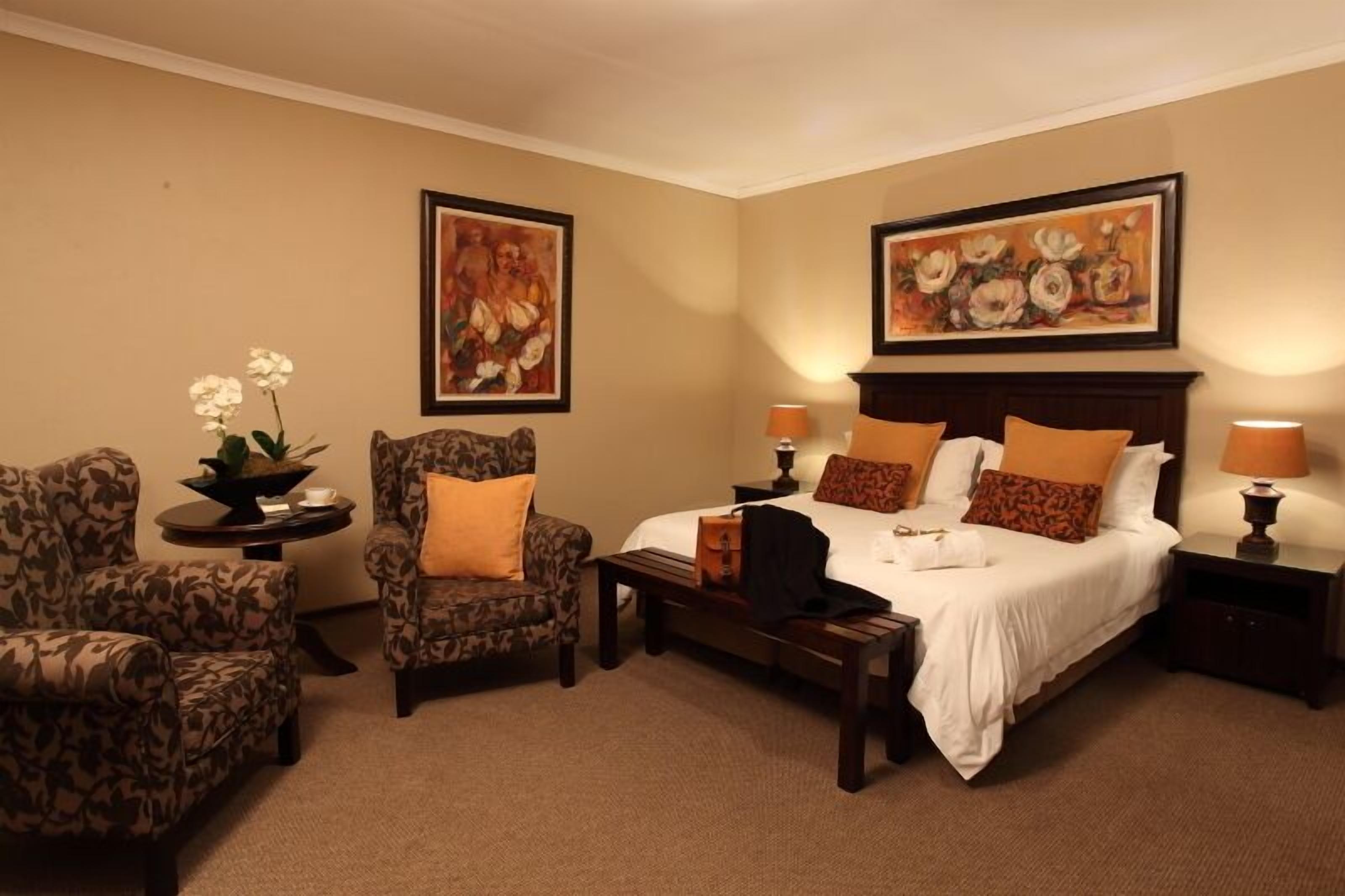 Quarto de hotel com uma cama, duas poltronas, uma mesinha e duas mesas de cabeceira. Há duas pinturas nas paredes e uma planta em vaso no lado esquerdo.