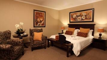 Quarto de hotel com uma cama, duas poltronas, uma mesinha e duas mesas de cabeceira. Há duas pinturas nas paredes e uma planta em vaso no lado esquerdo.