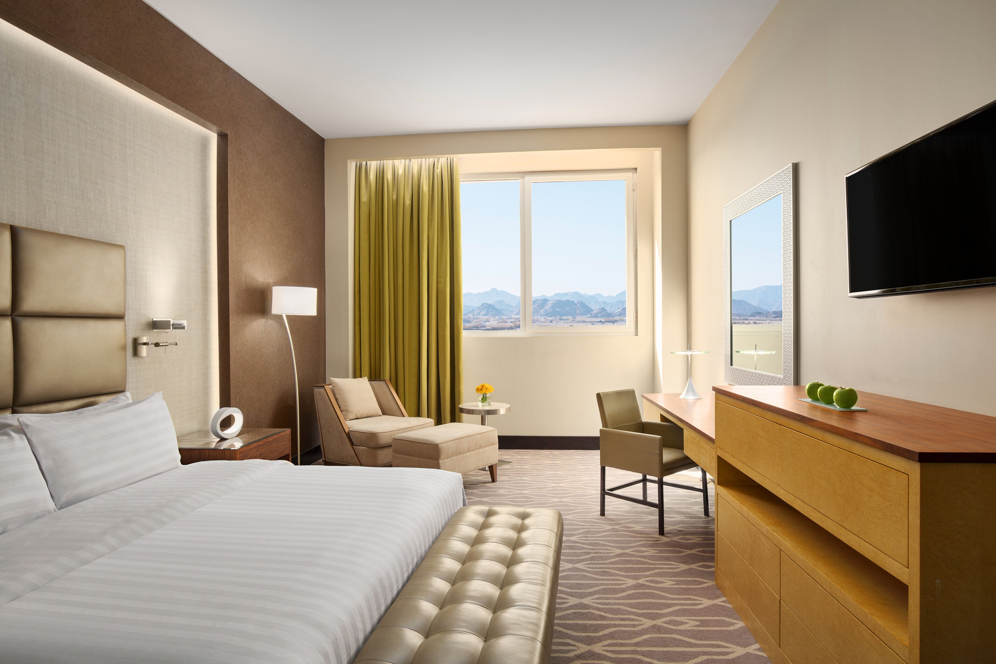 A hotel room with a bed, a desk, a chair, a TV, and a window with a view of mountains.