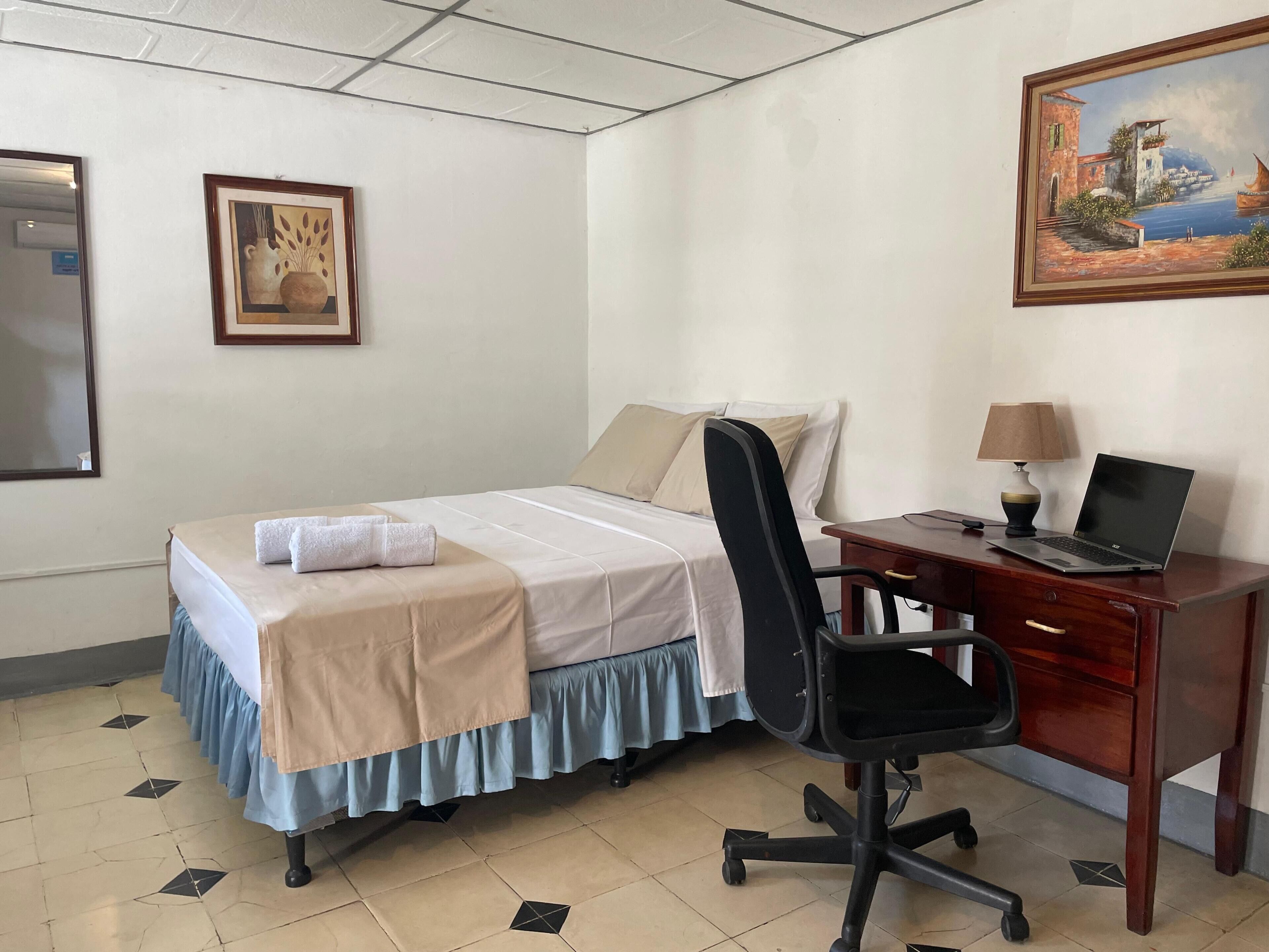Habitación de hotel con cama, escritorio con una computadora portátil, una silla, un cuadro en la pared y una fotografía enmarcada.