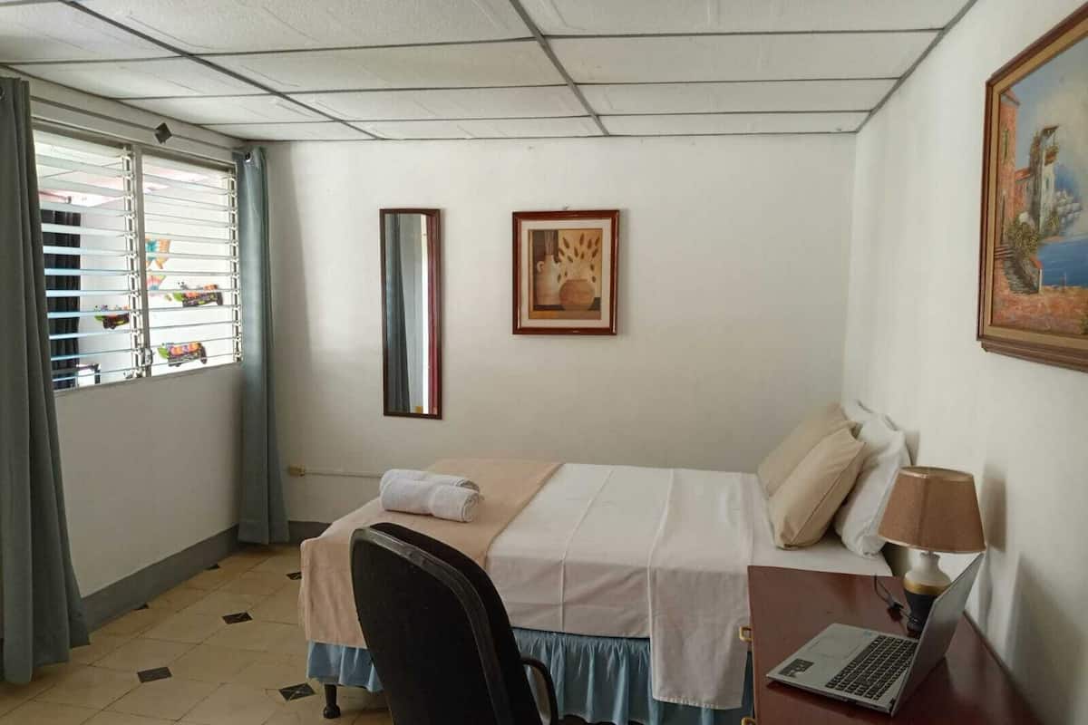 Hotelkamer met een bed, een bureau met een laptop, een stoel, een raam met jaloezieën en een schilderij aan de muur.