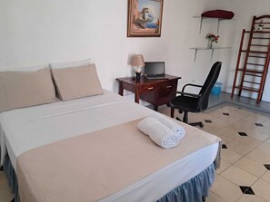 A neatly made bed with a rolled towel, a desk with a laptop, a chair, a small shelf with flowers, and a ladder against the wall. - El Callejon Guest House (León)