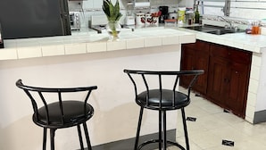Una cocina con dos taburetes negros, un refrigerador, un lavamanos y varios electrodomésticos.