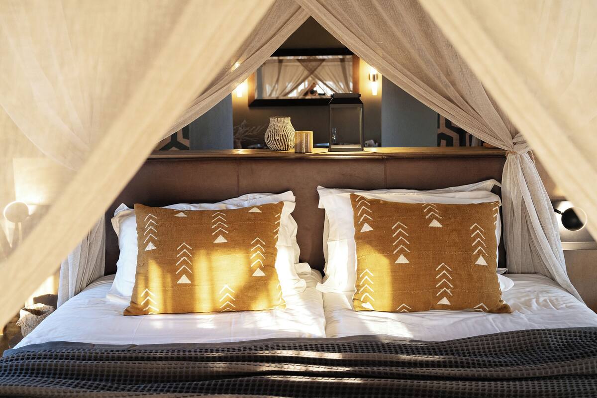 A bed with a canopy, pillows with a pattern, and a bedside table with a vase and a lantern.