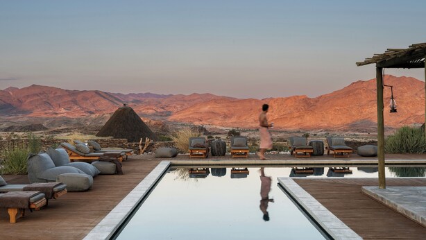 Un espace aménagé au bord de la piscine, avec des chaises longues et une structure dotée d’un toit de chaume, le tout surplombé par un paysage montagneux.