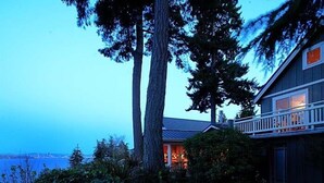 A house with a large tree in the front yard, a balcony on the right side, and a view of the water in the background. - Skiff Point Guest House (Bainbridge Island)