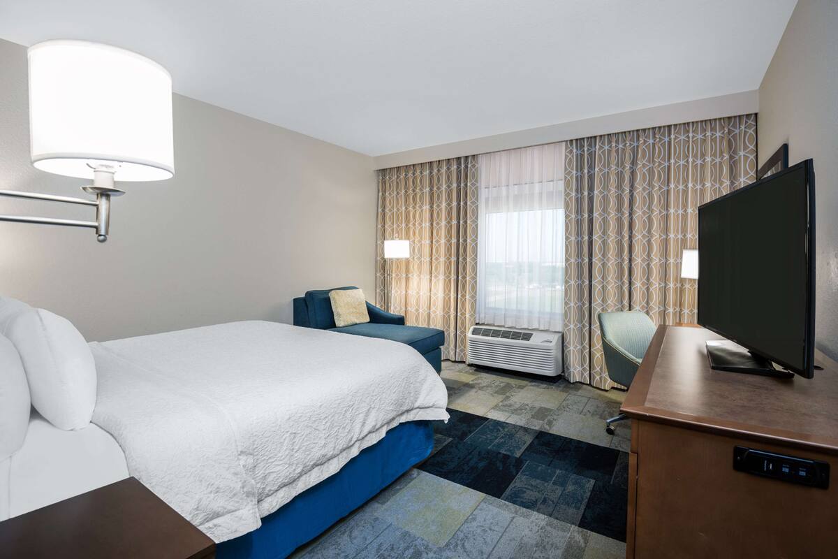 Une chambre d’hôtel avec un grand lit, un bureau avec une télévision, une chaise et une fenêtre avec des rideaux.