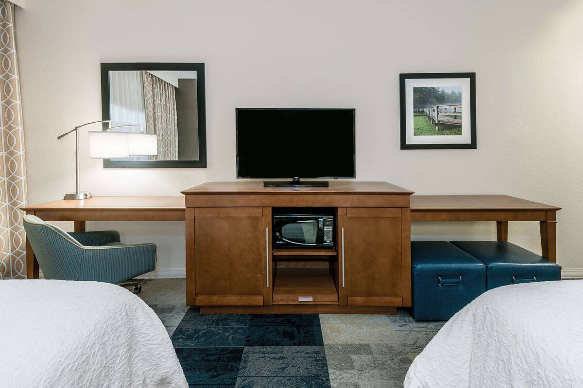 Une chambre d’hôtel avec un bureau en bois, une télévision, un four à micro-ondes, une valise bleue, deux lits, une chaise et une lampe.