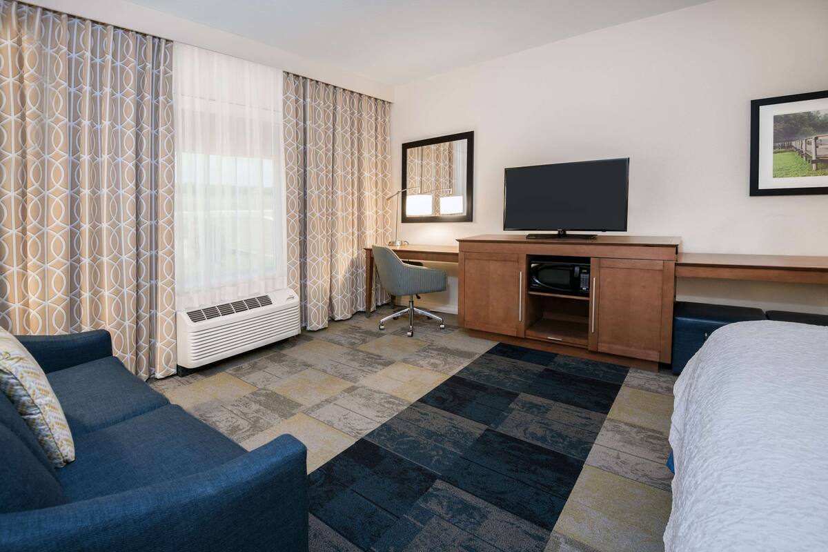 Une chambre d’hôtel avec un lit, un bureau avec une télévision, une chaise et une grande fenêtre avec des rideaux à motifs.