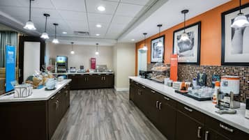 A modern kitchen with dark brown cabinets, white countertops, and a tiled backsplash. There are hanging pendant lights, a coffee machine, and a digital screen displaying a menu.