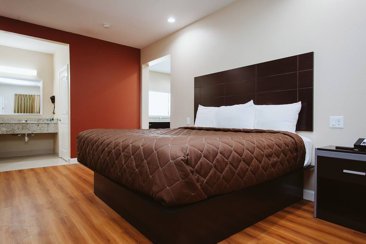 A modern hotel room with a large bed, wooden flooring, a built-in desk, and a bathroom visible through an open door.