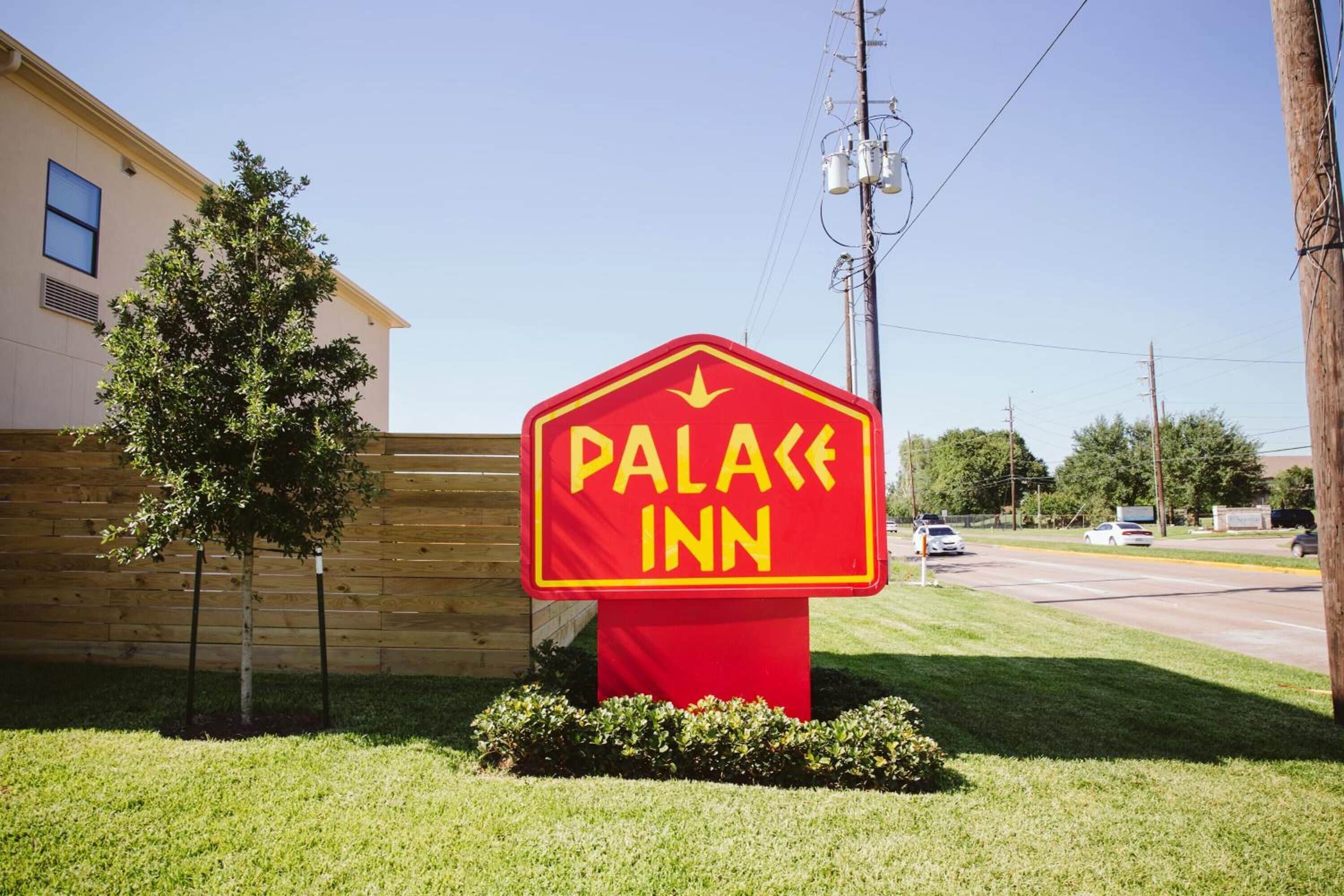 Señal para un hotel llamado "Palace Inn", con un diseño rojo y amarillo, ubicado frente a un edificio con valla de madera y un árbol.