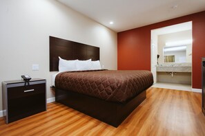 A hotel room with a large bed, a bedside table with a phone, a wooden floor, and a bathroom visible through an open doorway.