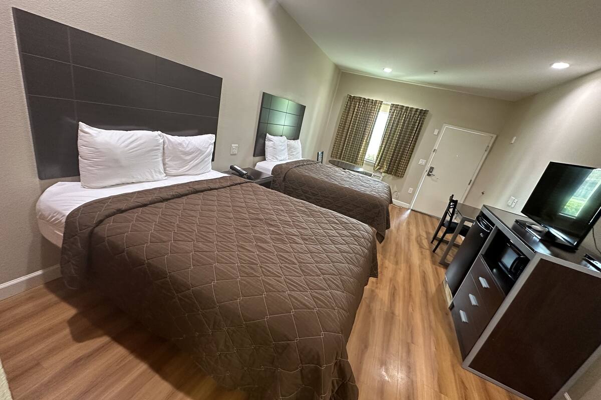 A hotel room with two beds, a TV, a kitchenette, and a bathroom.