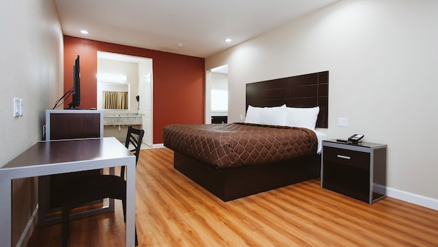A modern bedroom with a wooden floor, a bed with a dark headboard, a bedside table with a phone, and a desk with a chair. - Palace Inn 290 & Jones Rd (Houston)