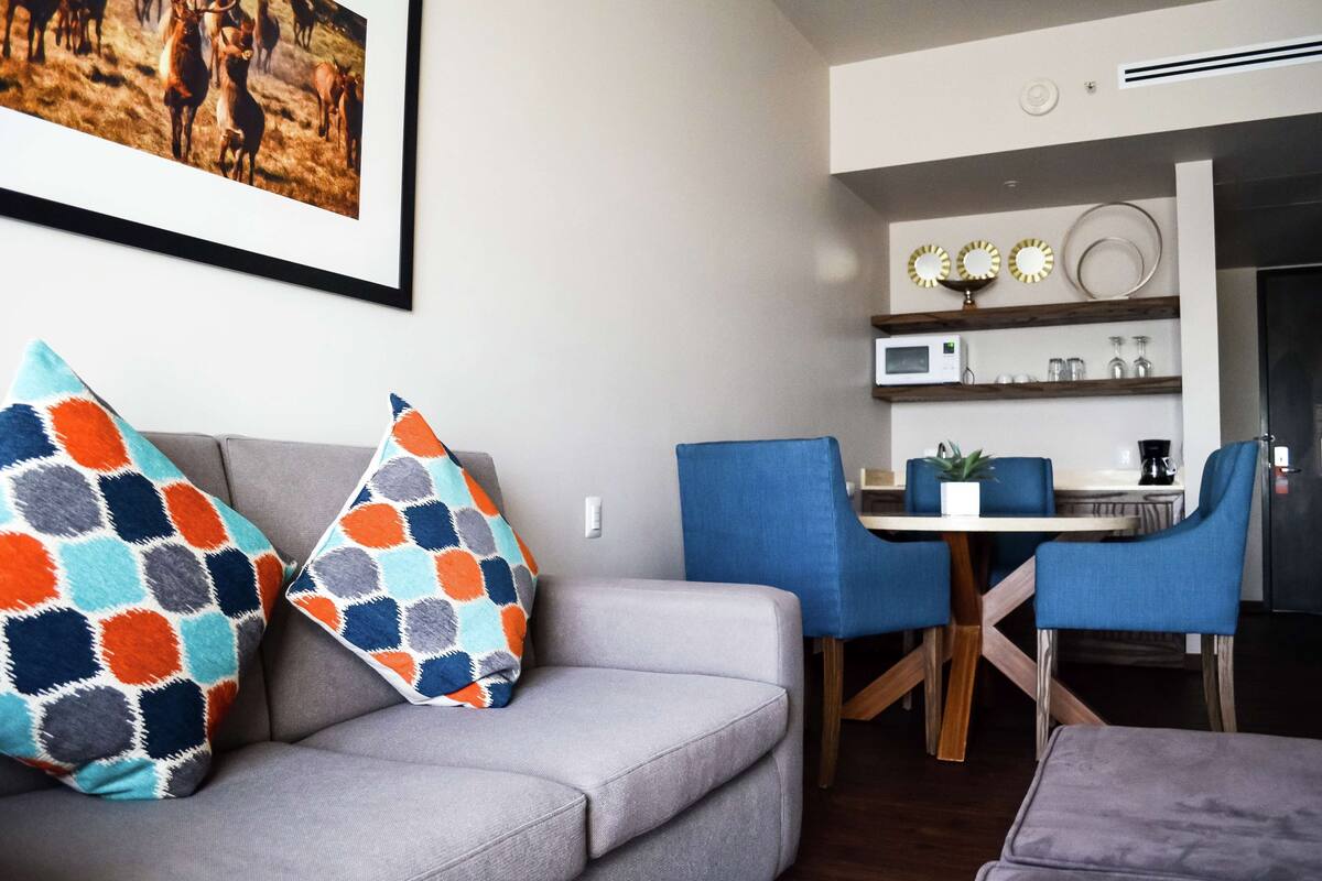 A modern living room with a grey sofa, colorful patterned pillows, a dining area with blue chairs, and a kitchen with a microwave and decorative items on the shelves.