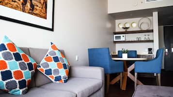 A modern living room with a grey sofa, colorful patterned pillows, a dining area with blue chairs, and a kitchen with a microwave and decorative items on the shelves.