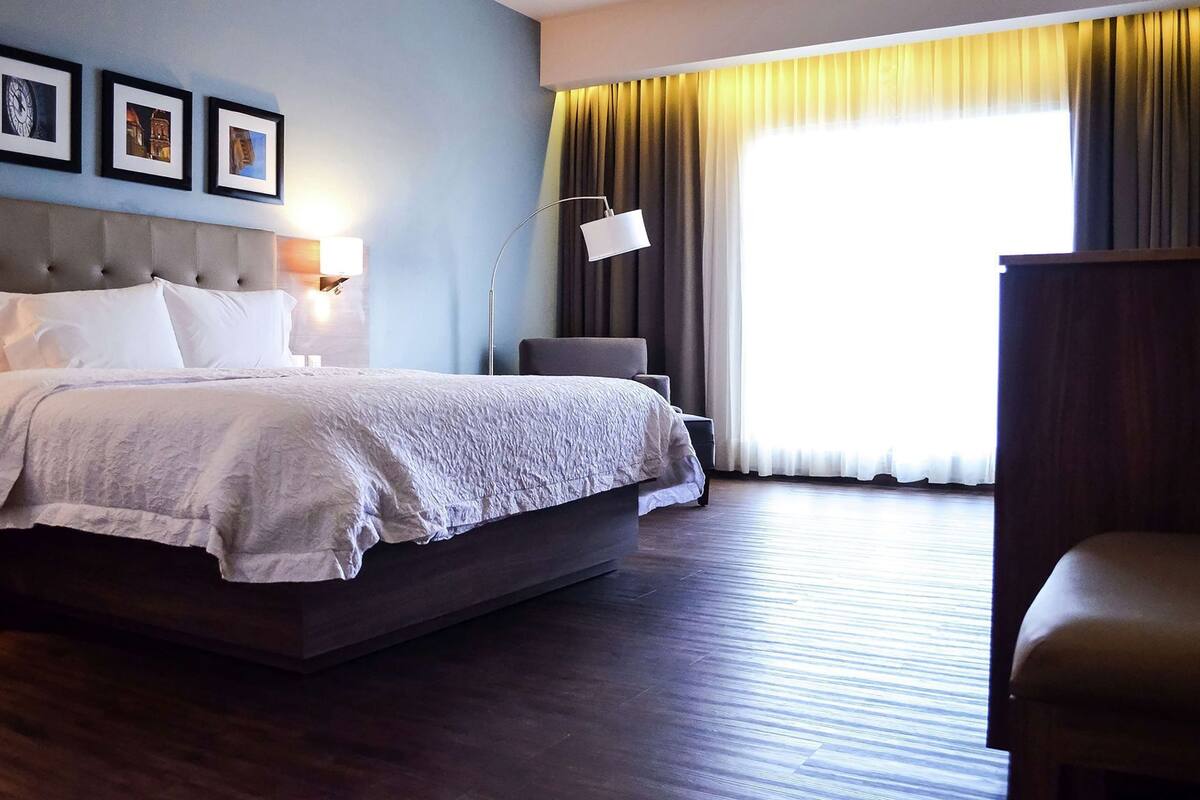 A hotel room with a large bed, a desk, a chair, a lamp, and a window with curtains.