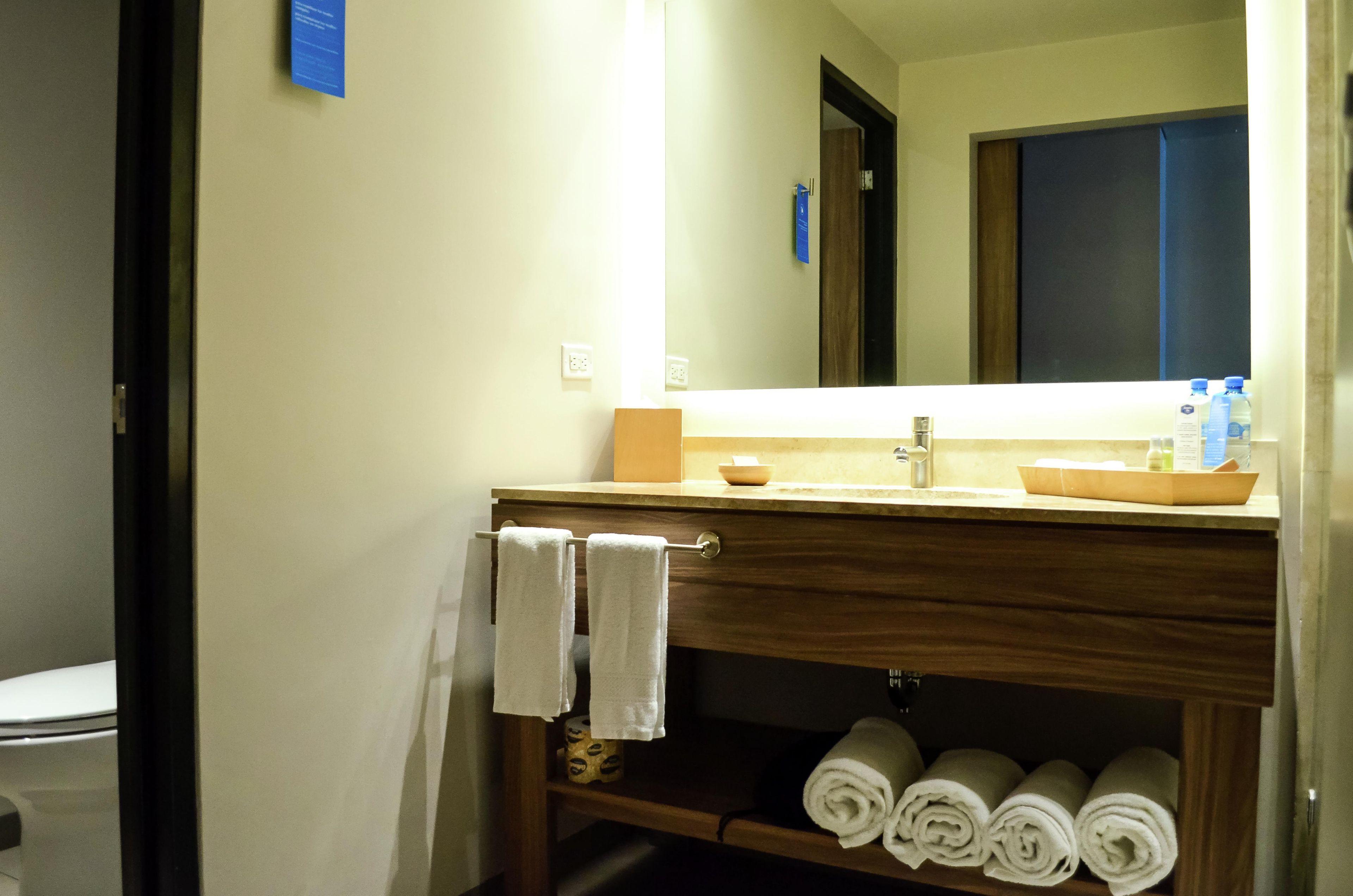 Un baño con lavamanos, espejo, toallero y repisa con toallas enrolladas.