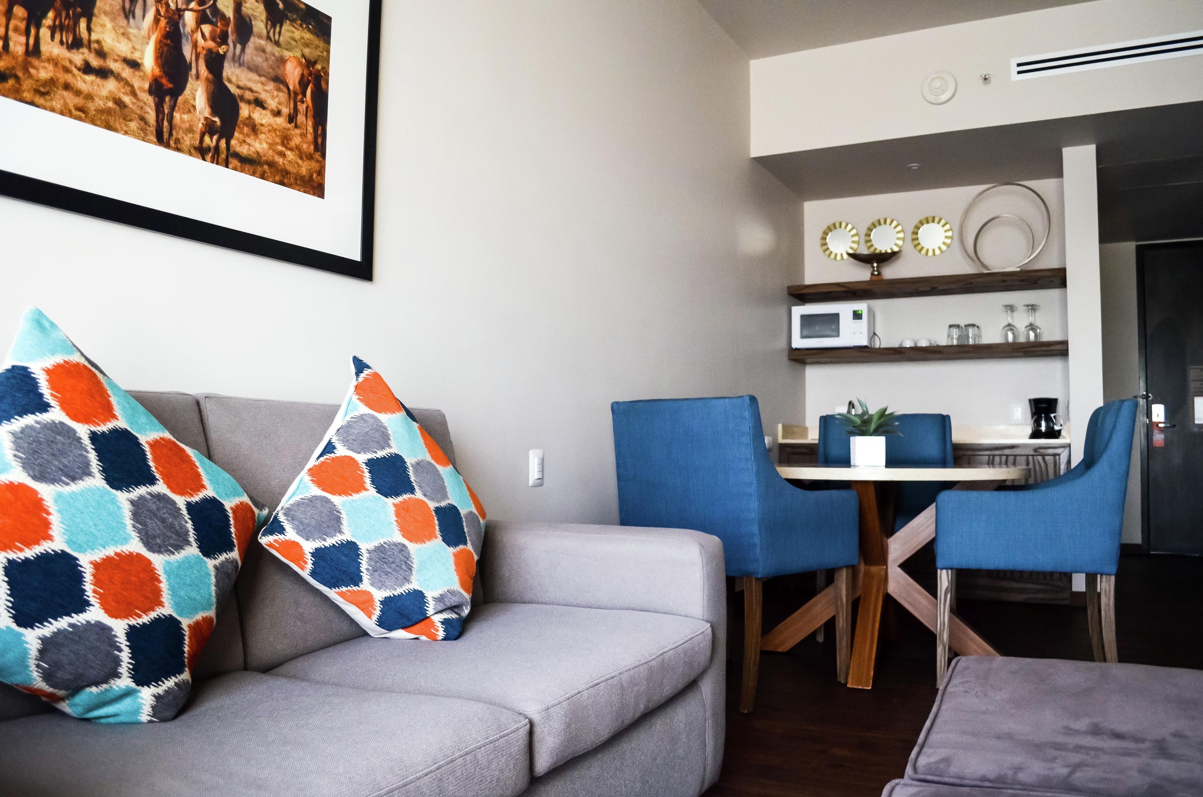 A living room with a grey sofa, two patterned throw pillows, a blue chair, a dining table with chairs, a microwave, and a small potted plant.