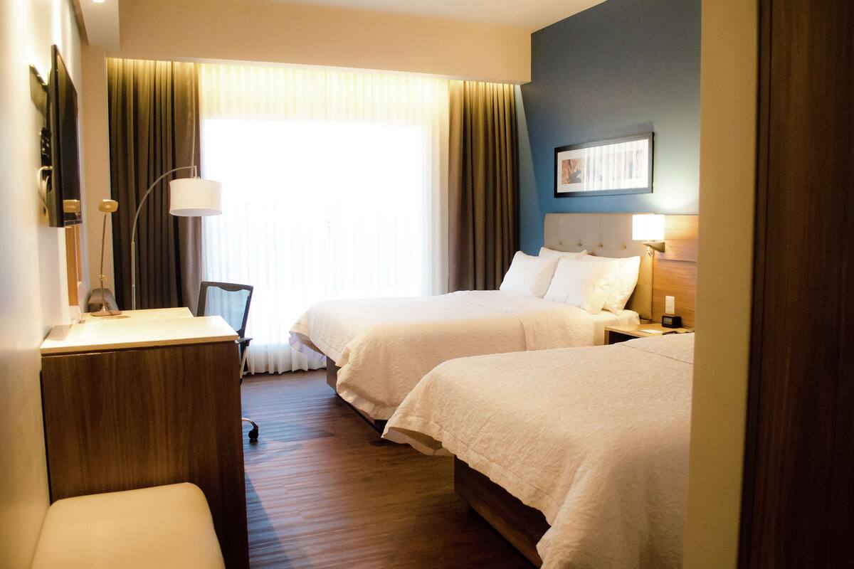 A hotel room with two beds, a desk, a chair, a lamp, and a window with curtains.
