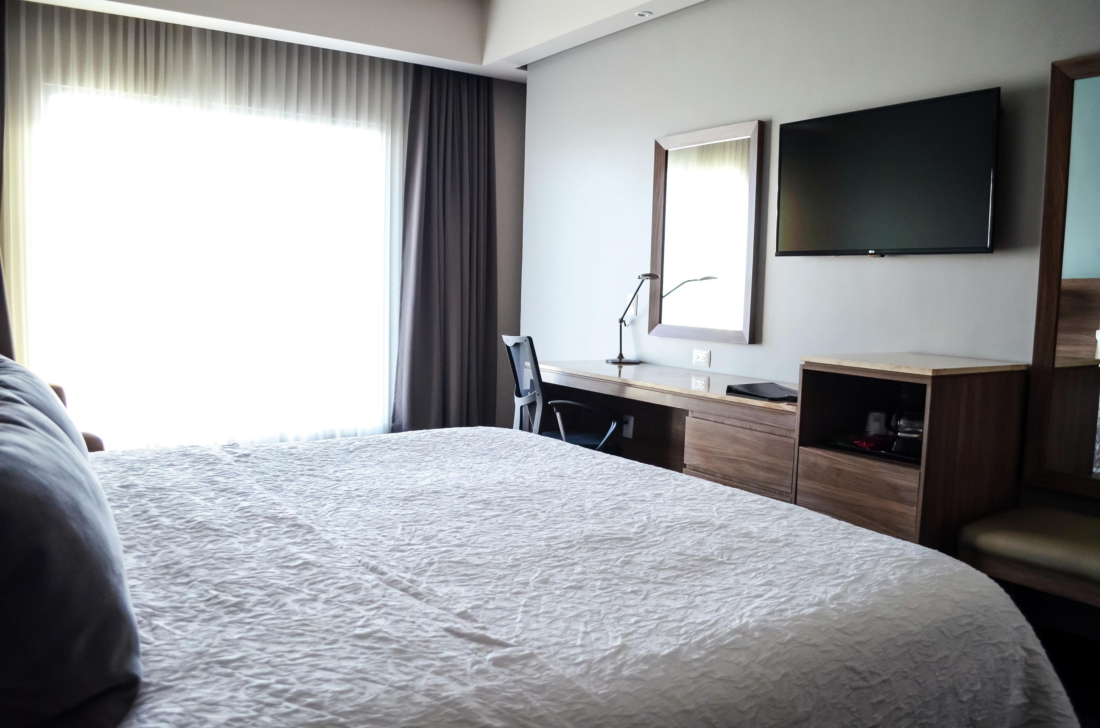 Une chambre d’hôtel avec un grand lit, un bureau avec une chaise, un téléviseur à écran plat fixé au mur et une fenêtre avec des rideaux.