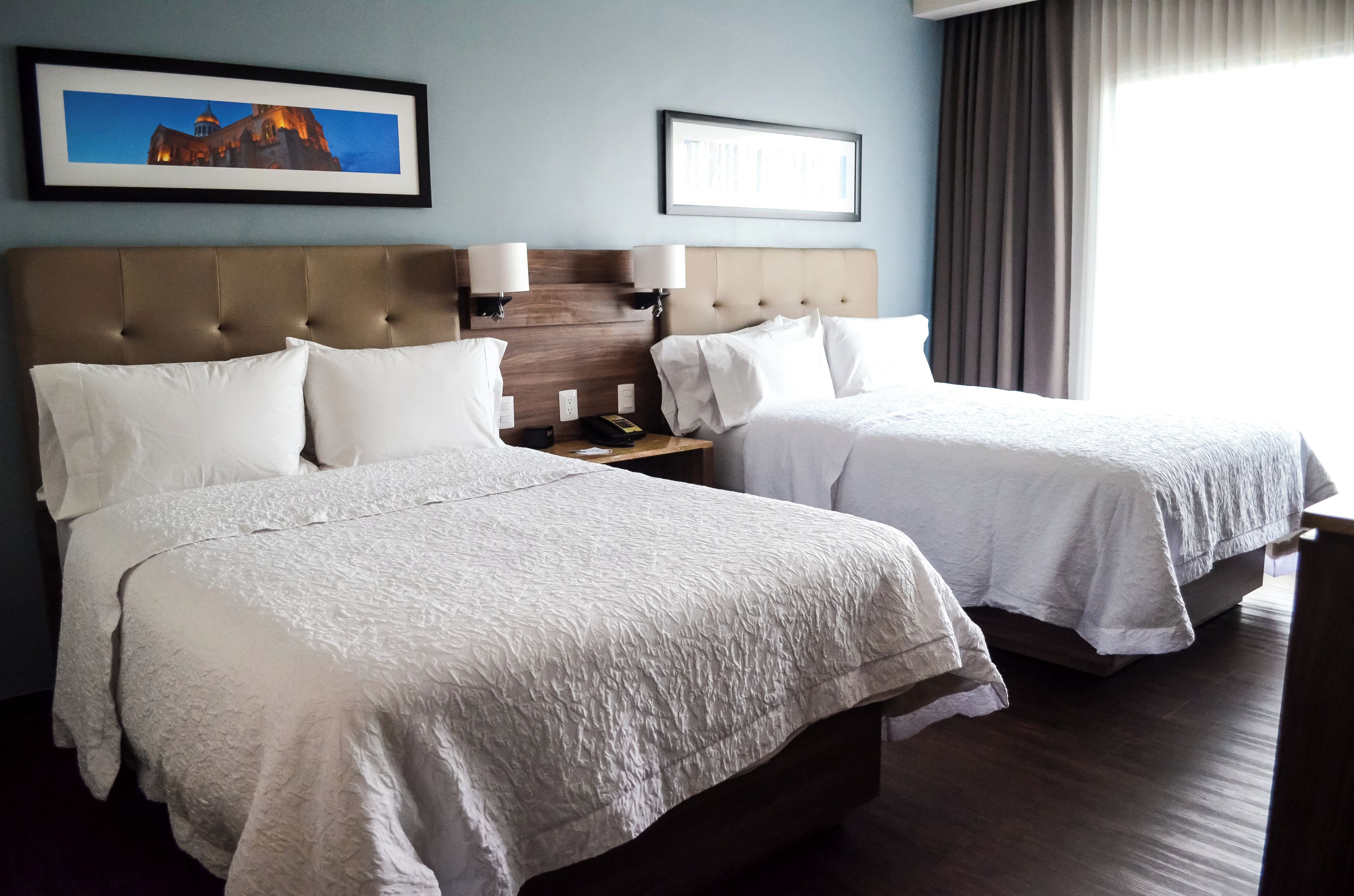 Une chambre d’hôtel avec deux lits, une tête de lit, des lampes de chevet, une table de chevet et un tableau encadré au mur.